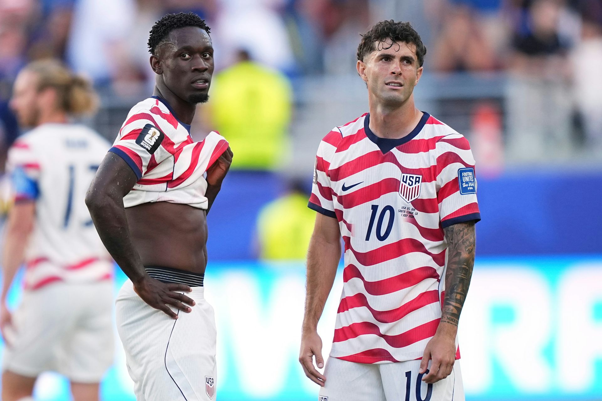 A black man and a white man in red and white striped jersey stand on a soccer field