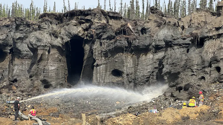 People work in reflective yellow/orange jackets at the base of a cliff, with a high-pressure jet of water spraying across