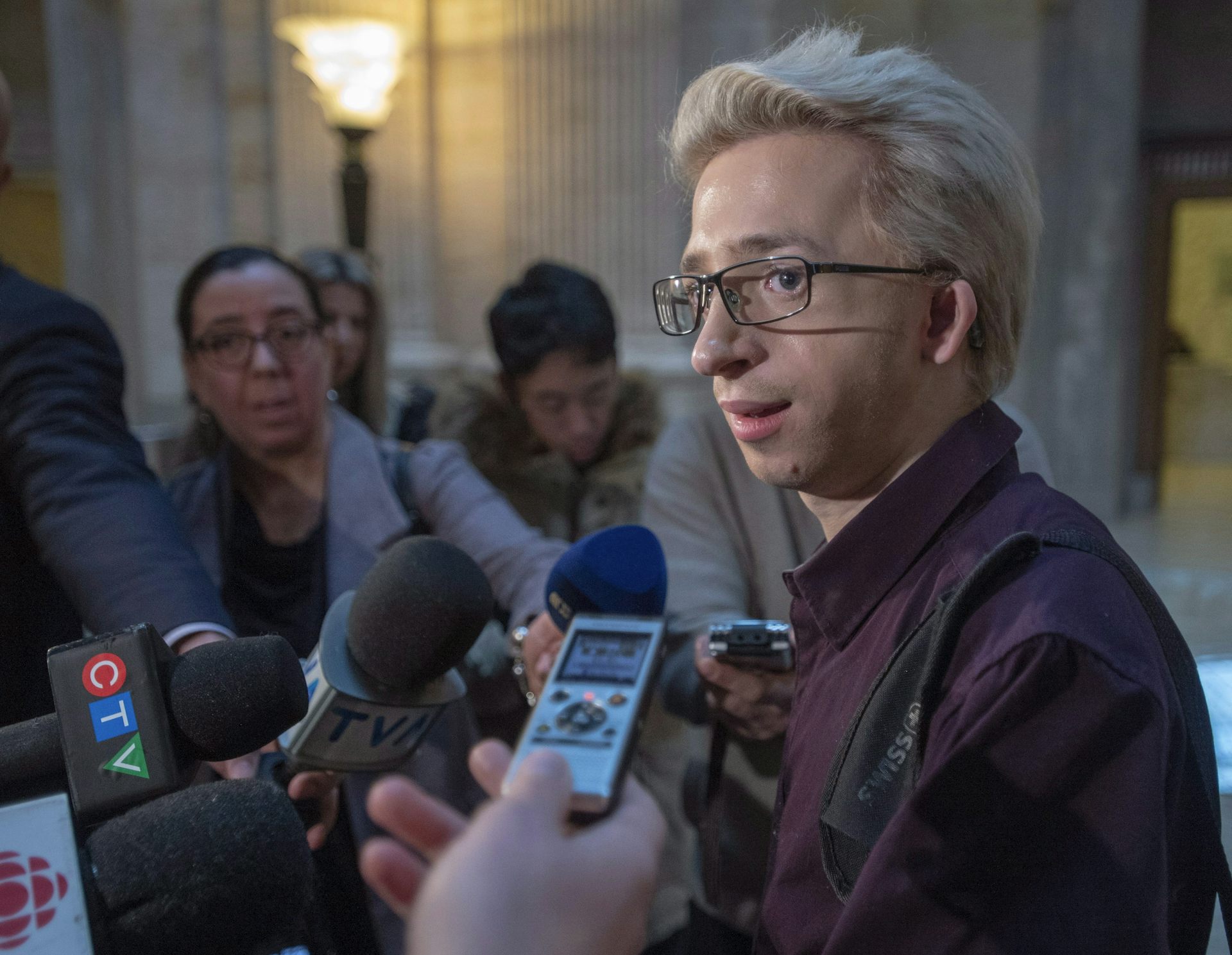 Un jeune homme aux cheveux blonds et portant des lunettes s'adresse aux médias.