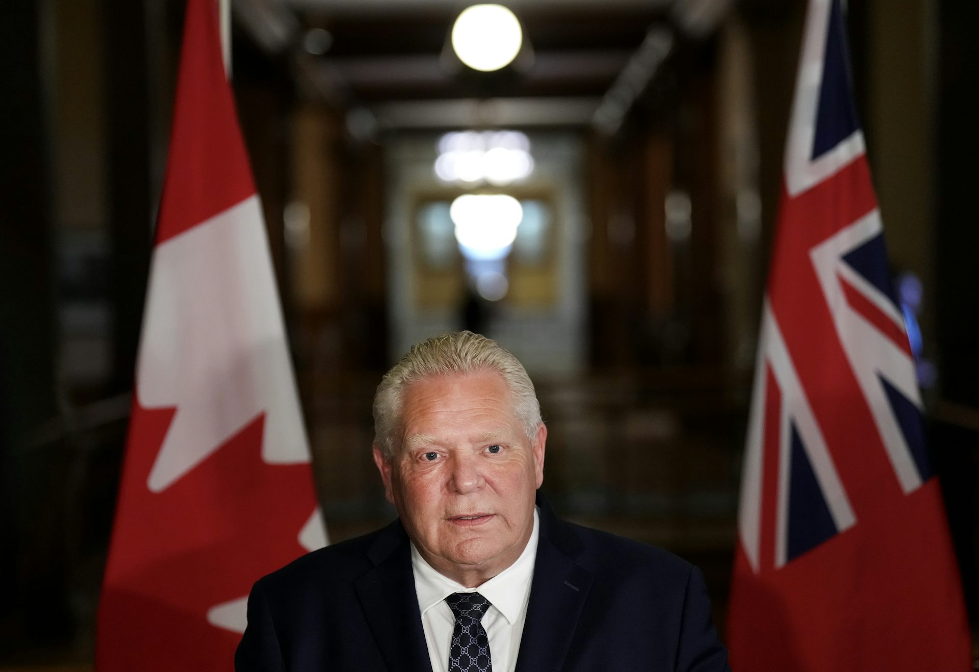 A rotund middle-aged white man stands in front of the Canadian flag and the Ontario flag