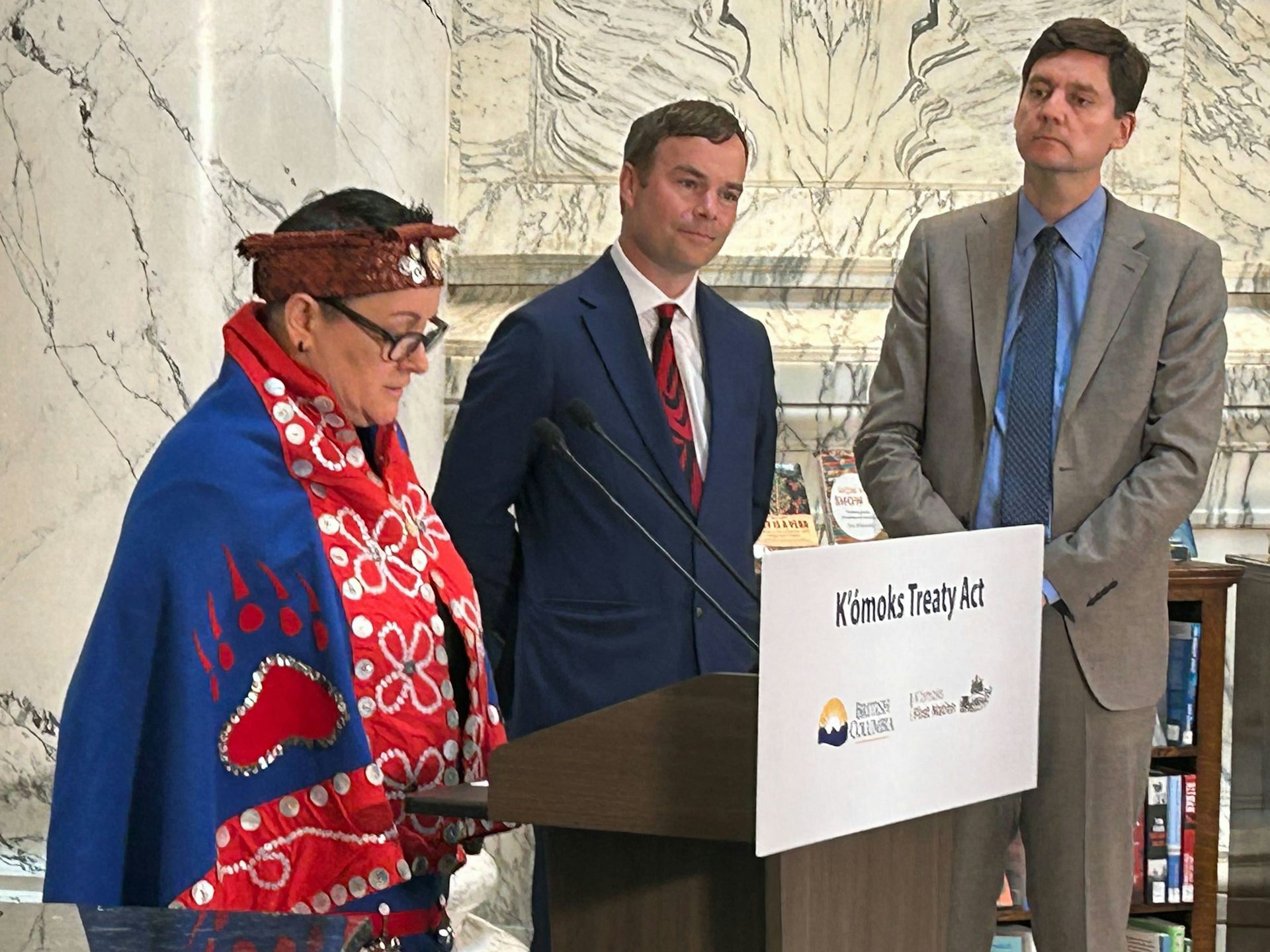 Two men in suits at a podium with a woman wrapped in a blanket with button design.