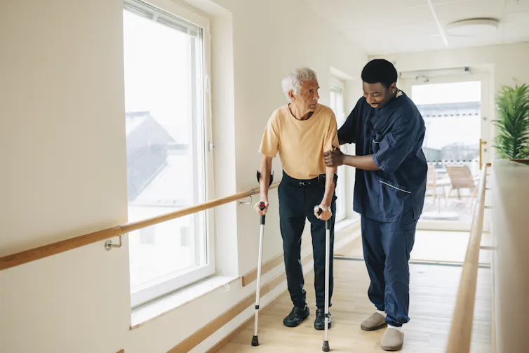 Male caregiver helping senior man walking with crutches in corridor at nursing home.