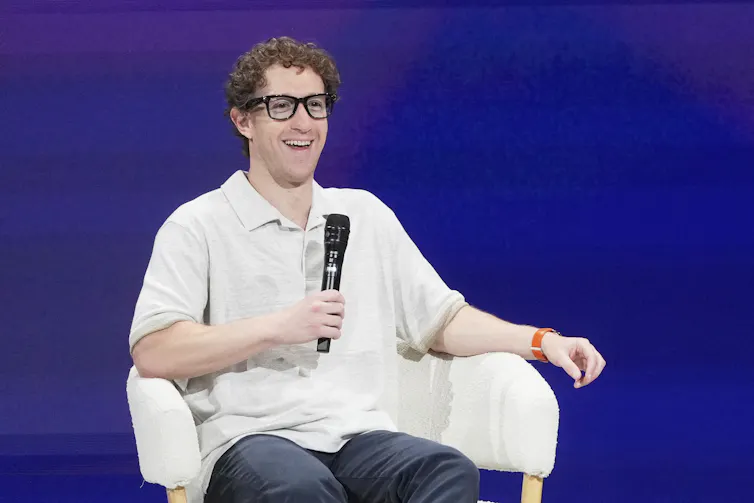 A middle aged man in glasses and white shirt sits in a chair with a microphone against a blue backdrop