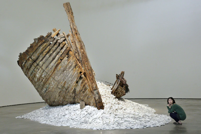 A woman squats next to a sculpture featuring a ship wreckage and pieces of porcelain.