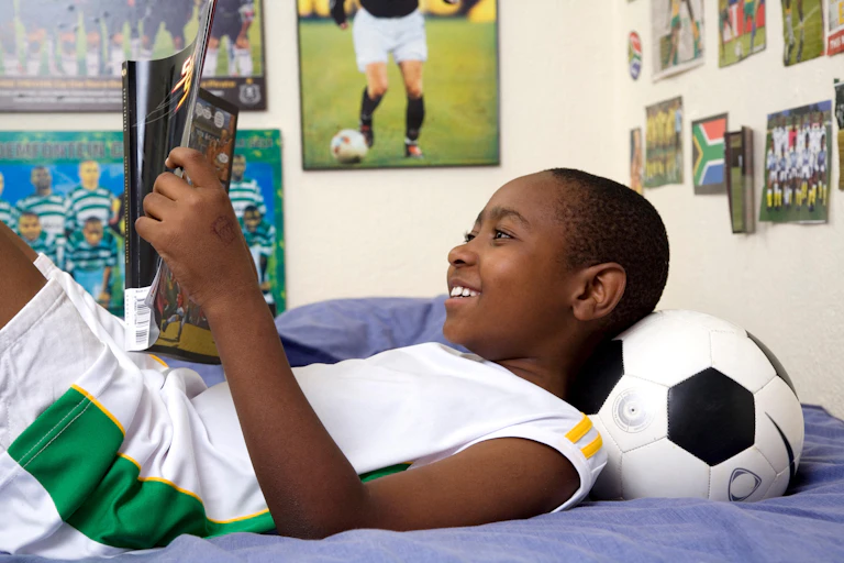A young boy on his bed surrounded by posters of football players reading a football magazine.