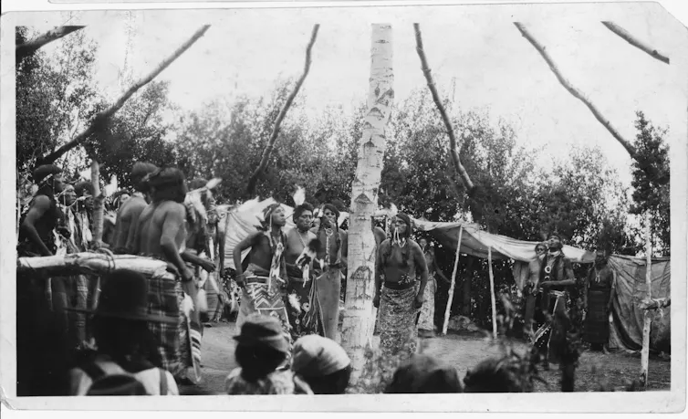 A black-and-white photo showing a ceremony in which several men are standing around a central tree trunk with polelike branches extending outward from it, while a crowd of people are gathered around.
