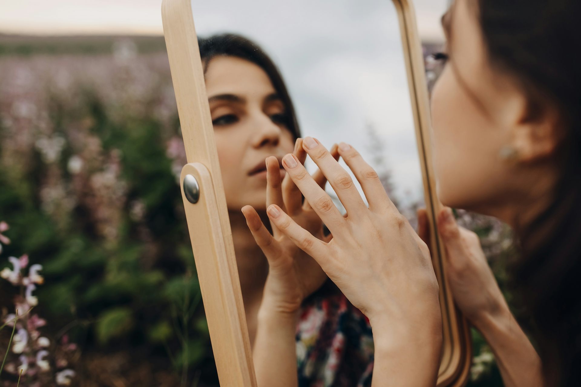 Woman stands in field touching her reflection in a mirror.