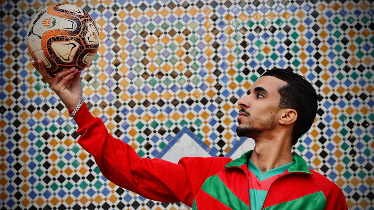 A young Moroccan man in a red and green football jersey holds a soccer ball aloft against a mosaic backdrop.