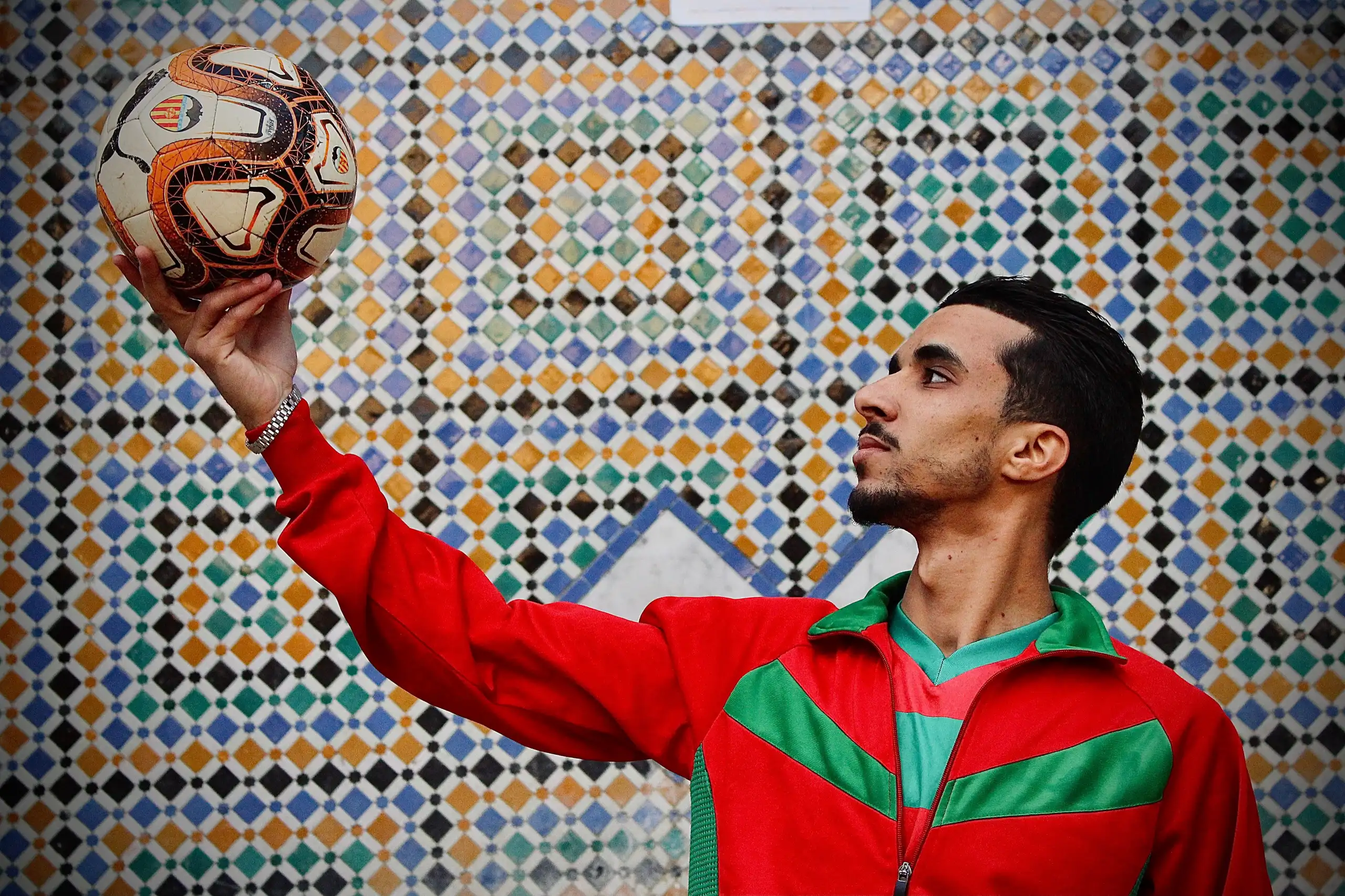 A young Moroccan man in a red and green football jersey holds a soccer ball aloft against a mosaic backdrop.