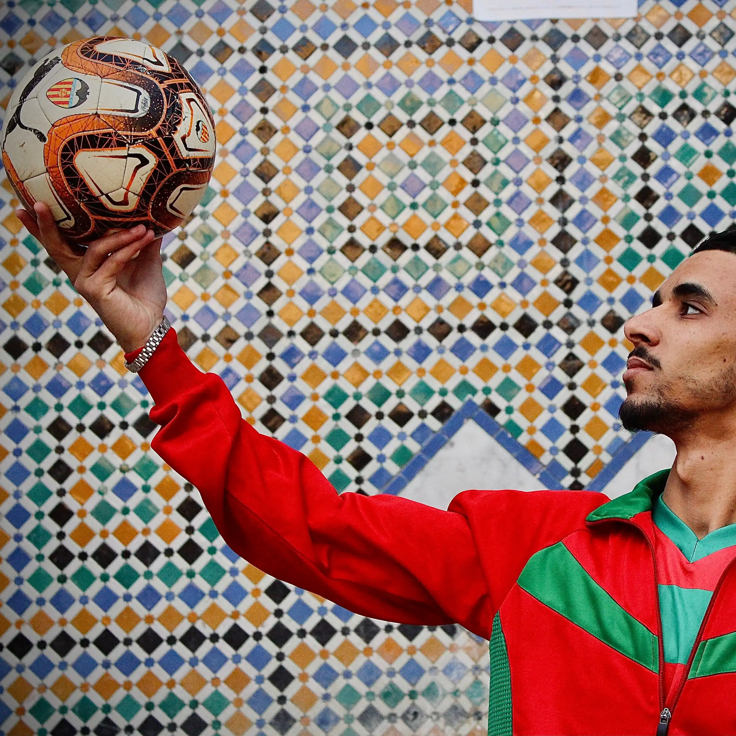 A young Moroccan man in a red and green football jersey holds a soccer ball aloft against a mosaic backdrop.