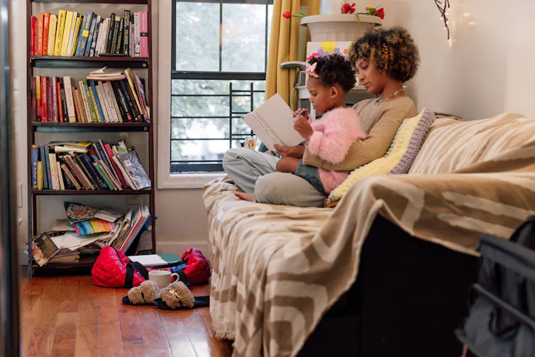 A woman sits on a couch and holds a small child in her lap as they both look at a book.