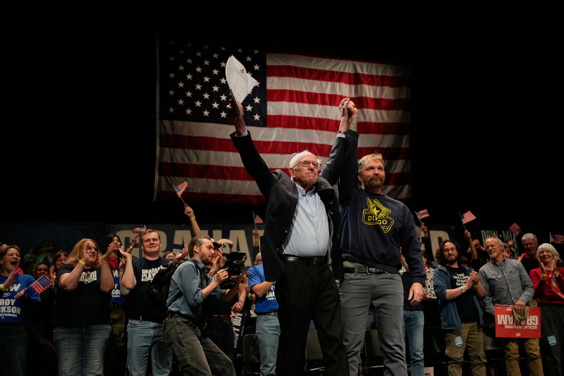 Two men on a stage backed by an American flag and a large group of people raise their arms together in triumph.
