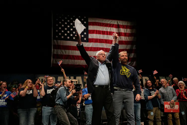 Two men on a stage backed by an American flag and a large group of people raise their arms together in triumph.