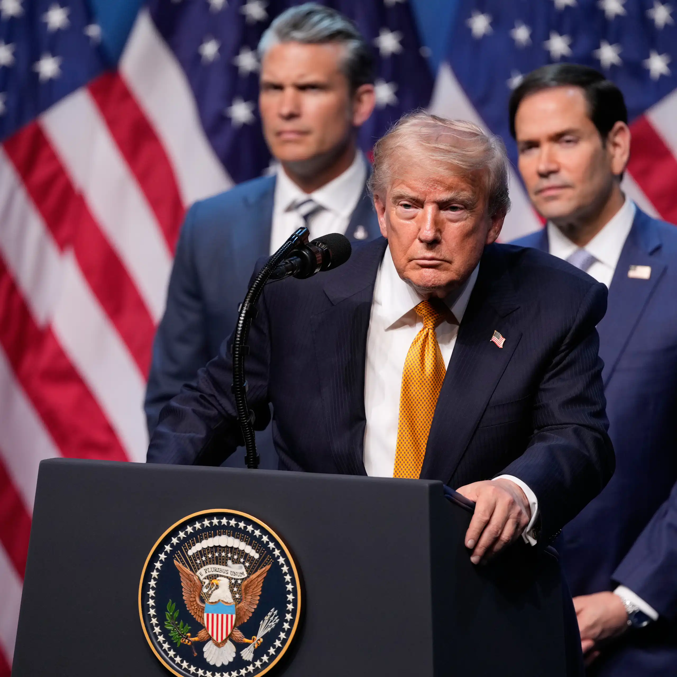 Donald Trump stands at lectern bearing presidential seal with Pete Hegseth, Marco Rubio and US flags behind him