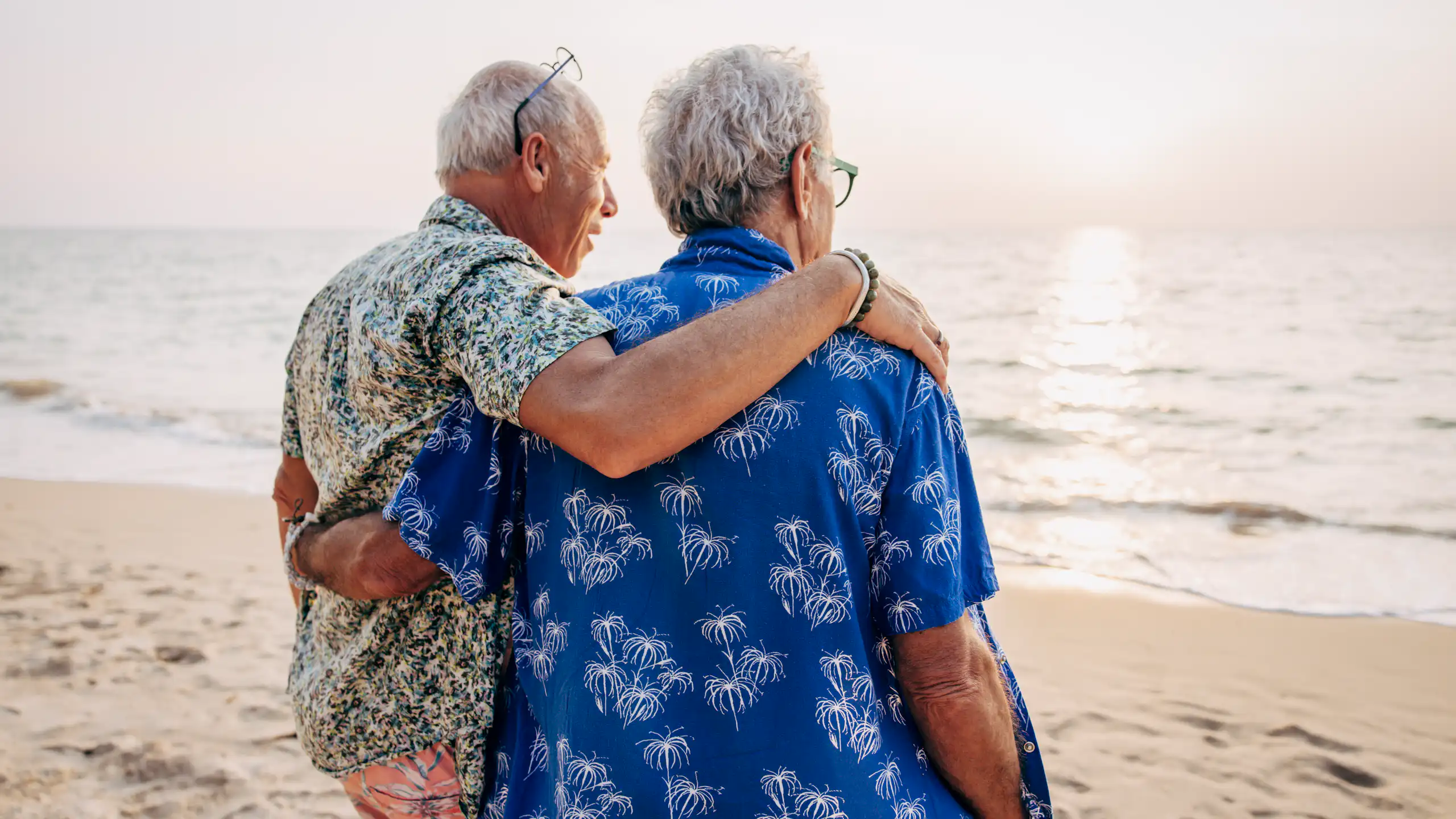 Two men with arms around each other, on a beach, looking out at the ocean