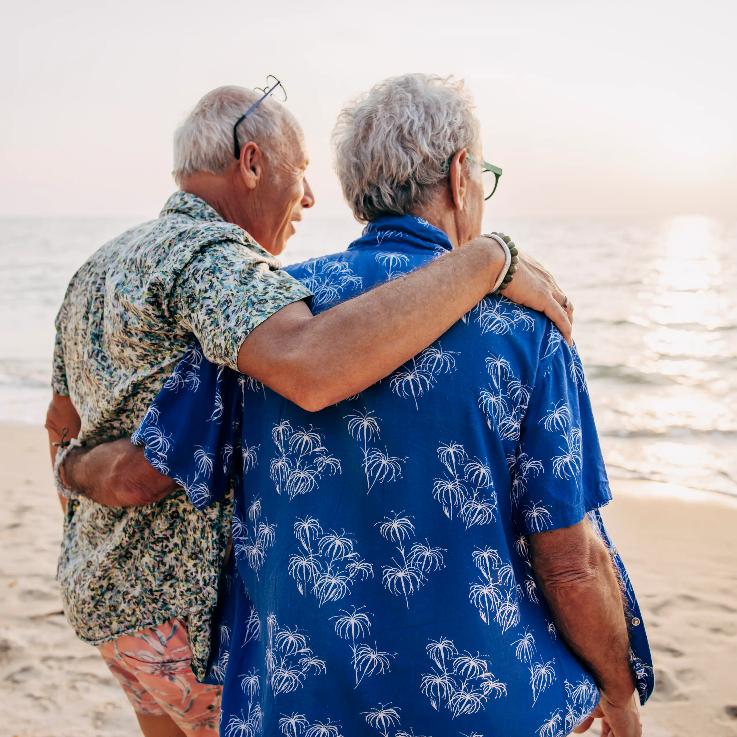 Two men with arms around each other, on a beach, looking out at the ocean