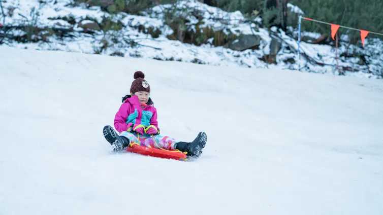 Young girl slides down a snowy mountain on a red toboggan.