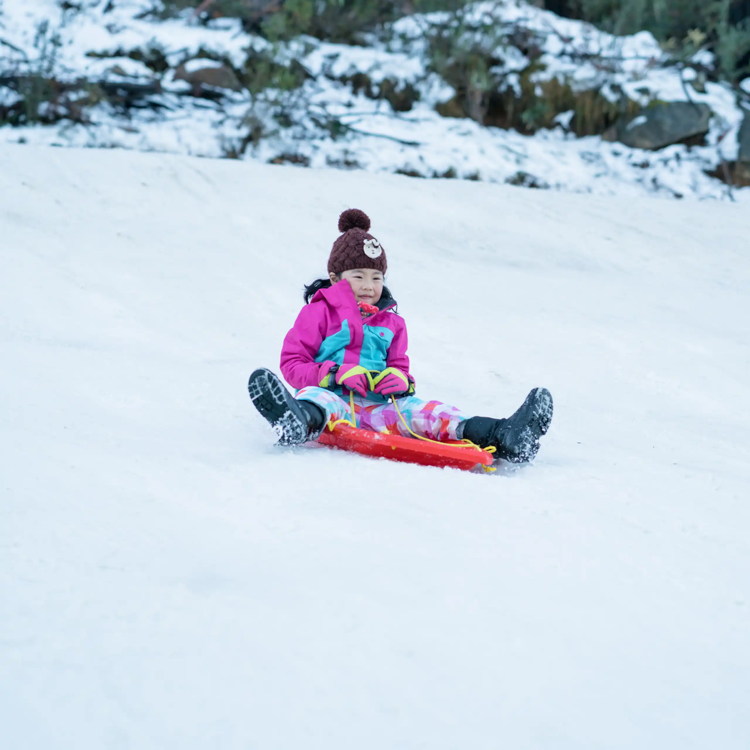 Young girl slides down a snowy mountain on a red toboggan.
