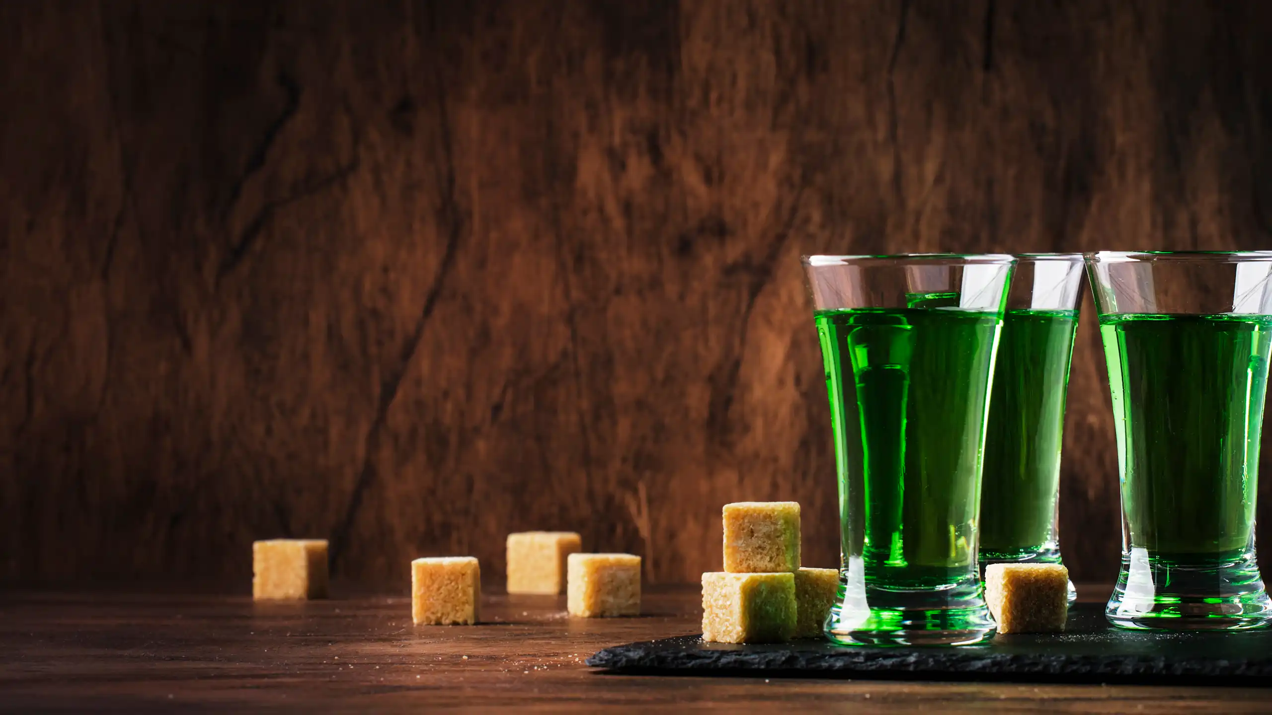 Série de verres d'absinthe au vert émeraude et de morceaux de sucre roux sur fond sombre couleur bois.