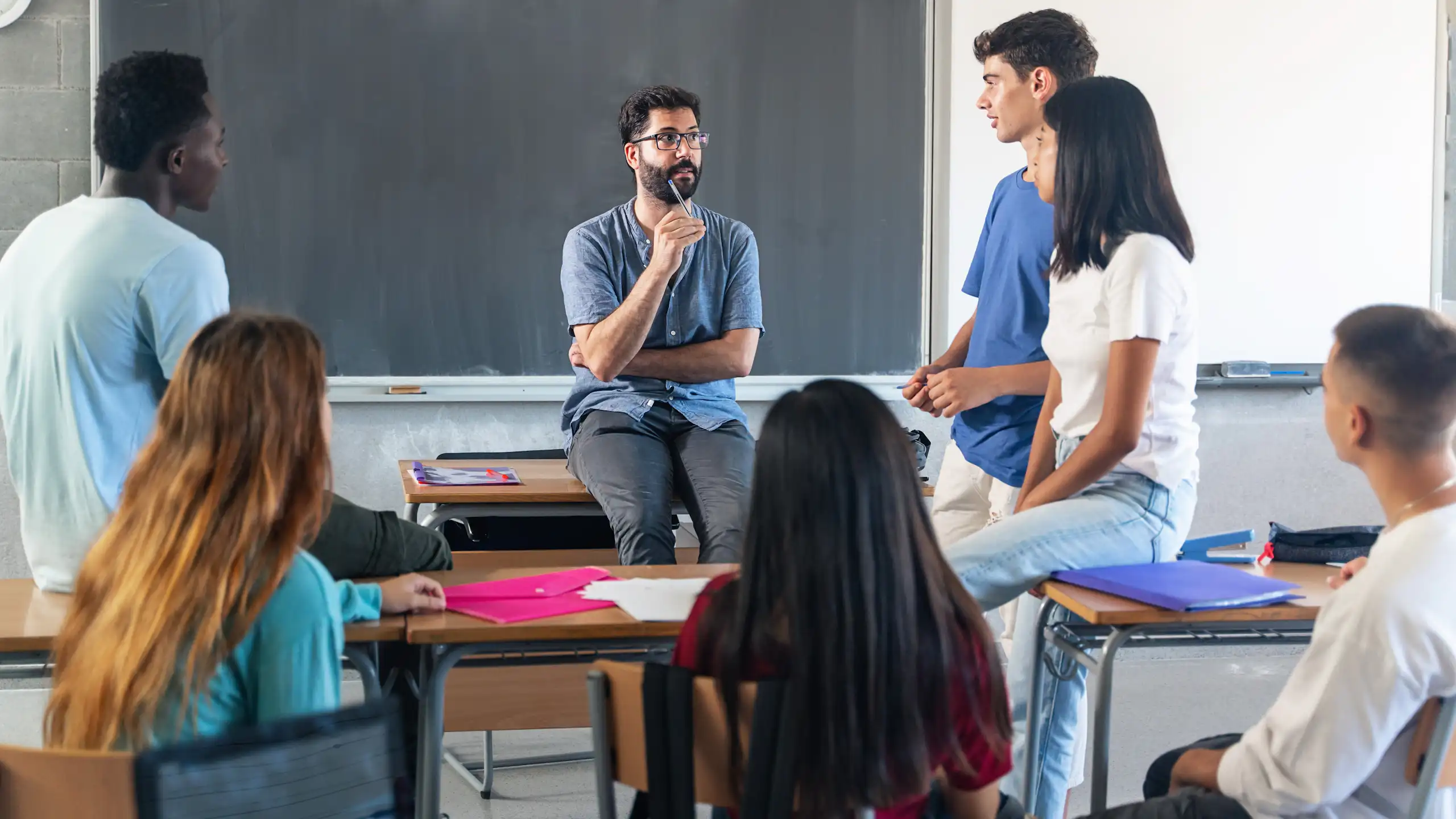 Un profesor explica en clase delante de la pizarra sentado en una mesa con varios alumnos de pie y sentados alrededor