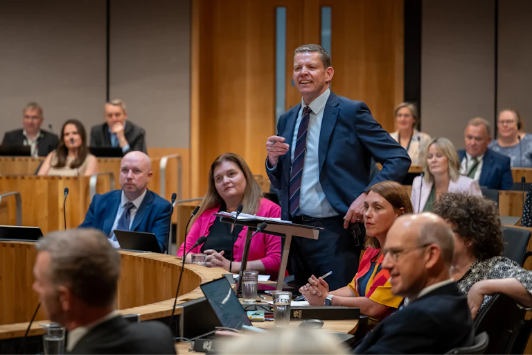 Rhun ap Iorwerth stands at the lectern in the Senedd chamber surrounded by other politicians.