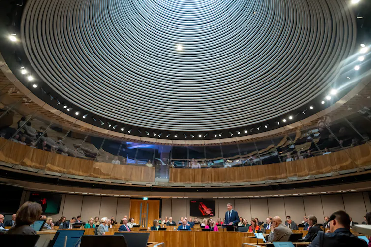 Wide shot of Rhun ap Iorwerth in the Senedd chamber surrounded by other politicians.