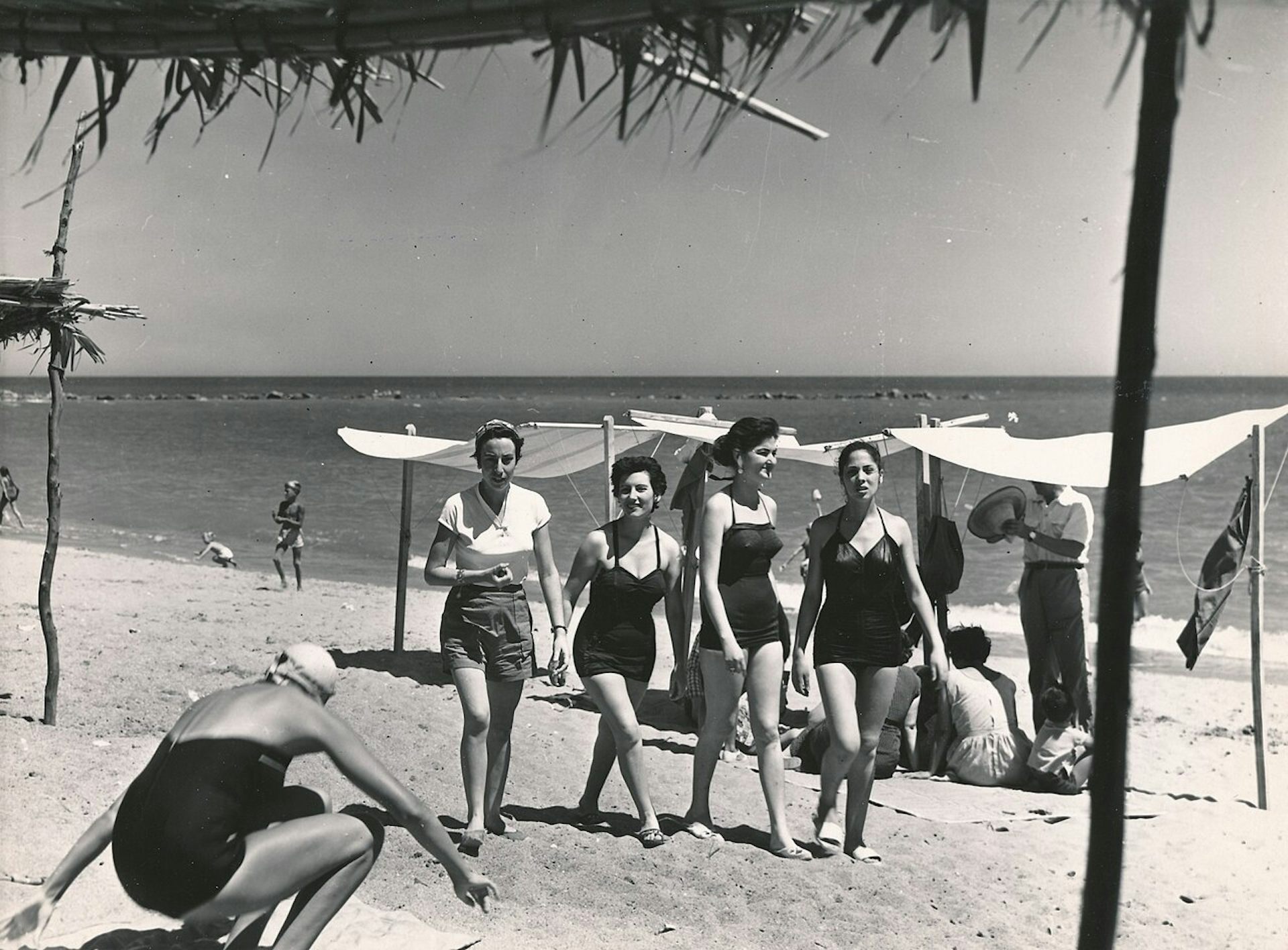 Fotografía en blanco y negro de cuatro chicas jóvenes en trajes de baño caminando por la playa.