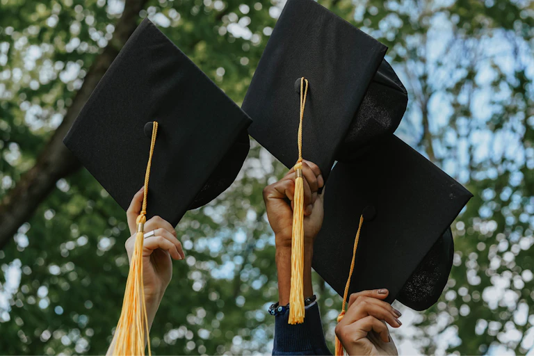Hands holding graduate caps up amid trees.