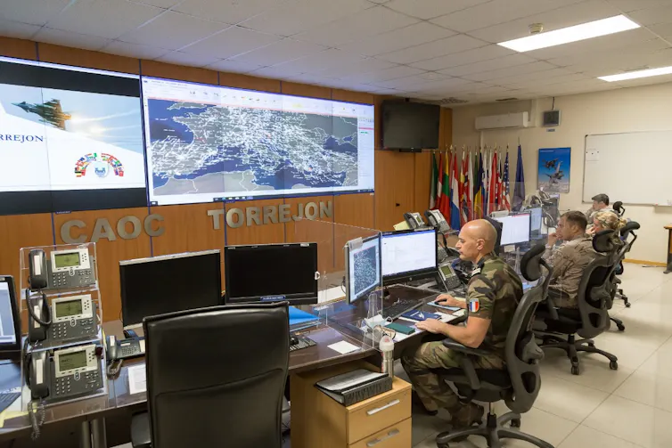 Militares sentados en sillas ante escritorios con pantallas y con una gran pantalla de pared que muestra vuelos en Europa