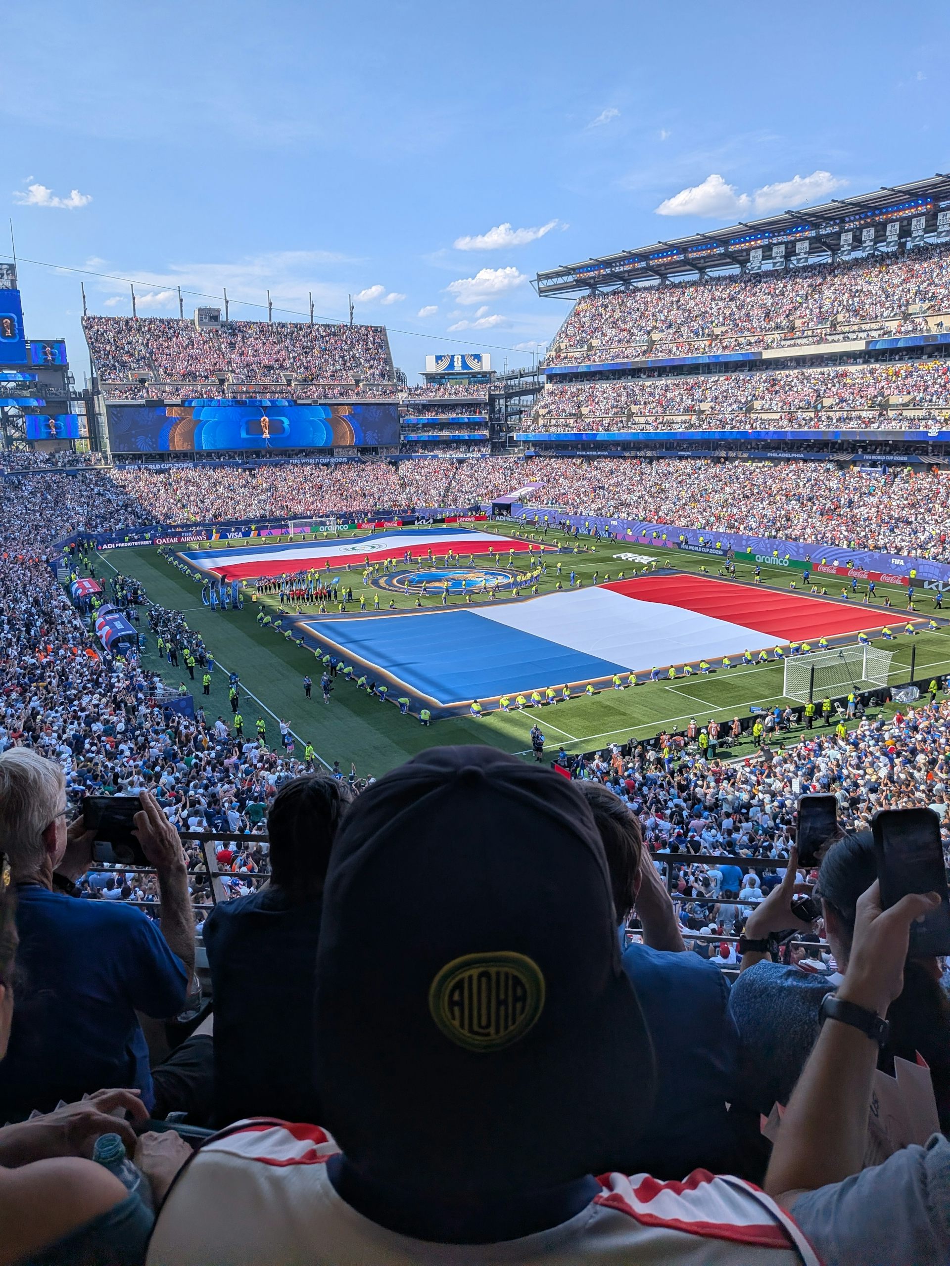The back of a person's head, wearing a cap, in a stadium full of people while giant flags are formed on the field.