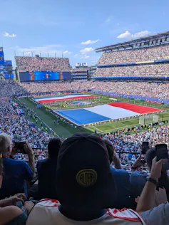 The back of a person's head, wearing a cap, in a stadium full of people while giant flags are formed on the field.
