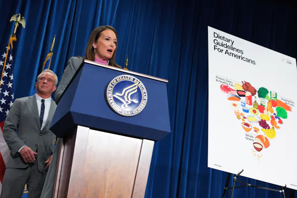 A woman speaks from behind a podium, a poster showing an inverted food pyramid on display nearby