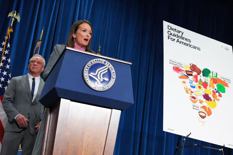 A woman speaks from behind a podium, a poster showing an inverted food pyramid on display nearby