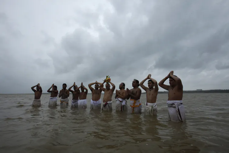 A line of around a dozen men wearing white sarongs stand in waist-high water, holding their hands up in prayer.