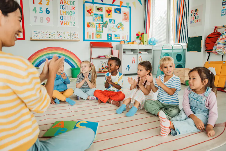 A row of young children sit on the floor, clapping, as they face a seated teacher in a striped yellow shirt.