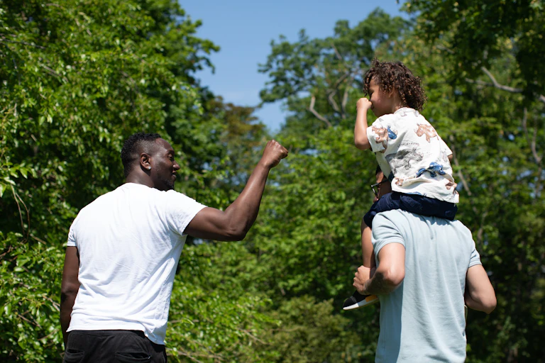 A child is seen riding on an adult's shoulders, and is giving a fist bump to another adult walking beside them.
