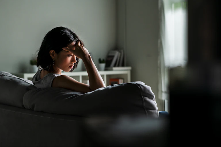 Young Asian woman sits on a sofa looking sad and worried.
