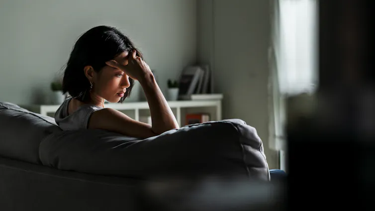 Young Asian woman sits on a sofa looking sad and worried.