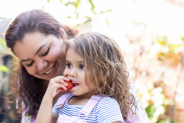 A mother holds her young daughter while the child eats a tomato