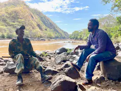 Two men sit on rocks with a lake and mountains in the background