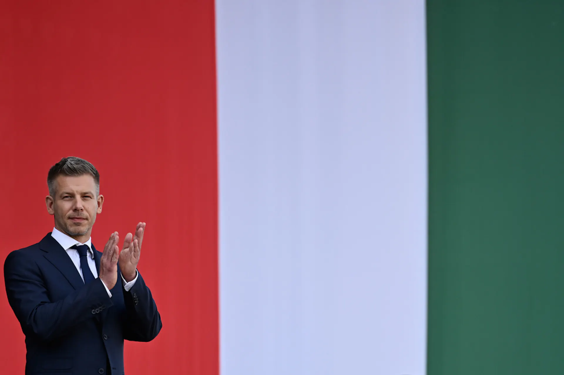 A man in a suit claps with a giant flag behind him.