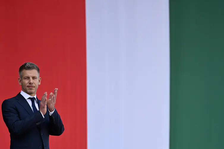 A man in a suit claps with a giant flag behind him.