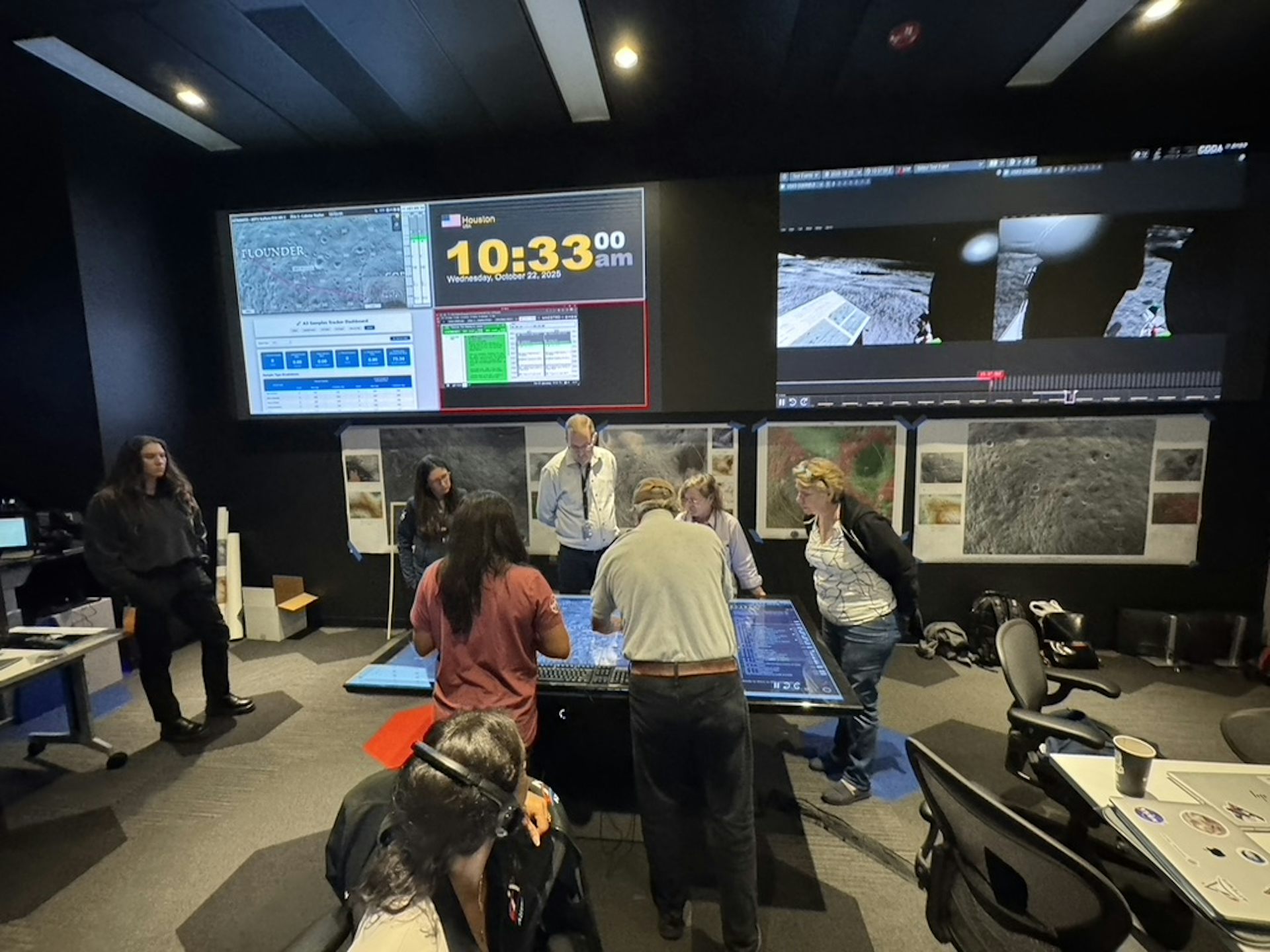 A group of people work together around a table in a room with large screens and equipment.