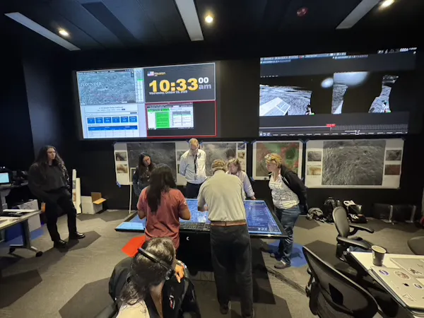 A group of people work together around a table in a room with large screens and equipment.