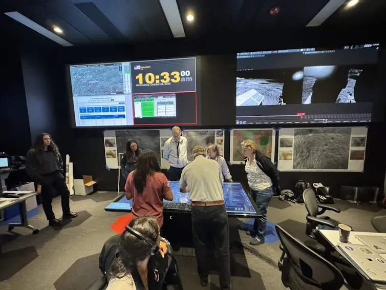 A group of people work together around a table in a room with large screens and equipment.