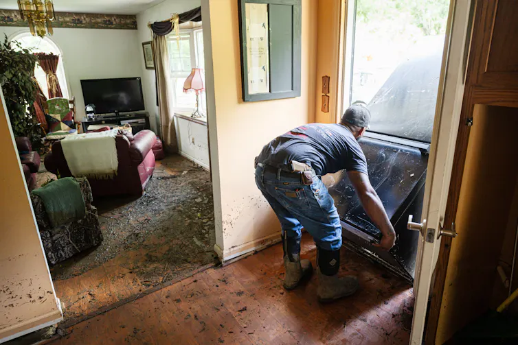 A man moves furniture in a home with mud covering the floor and parts of the walls.