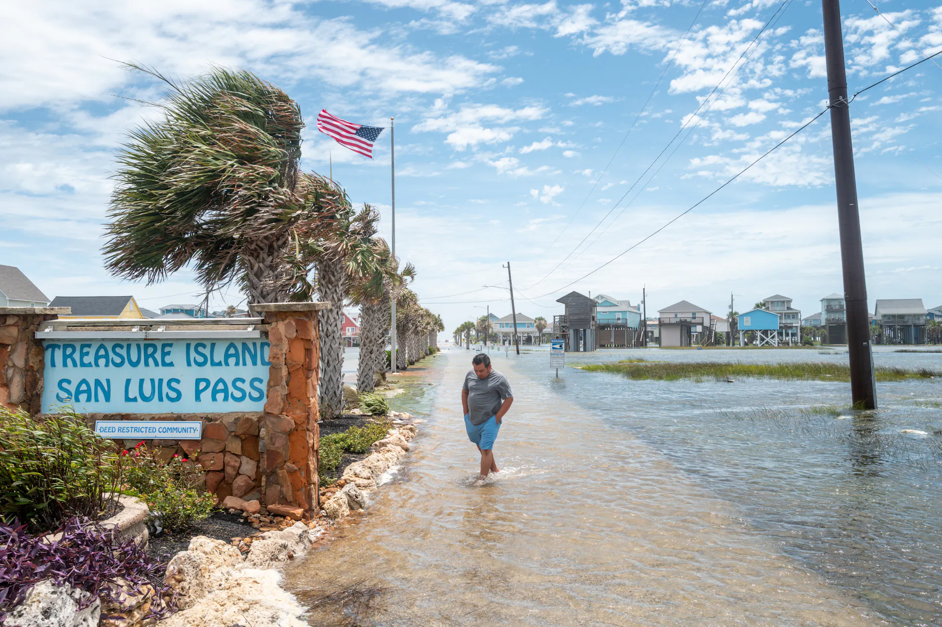 A man walks through ankle-deep water along a street beside homes. Other homes in the background are built on stilts.