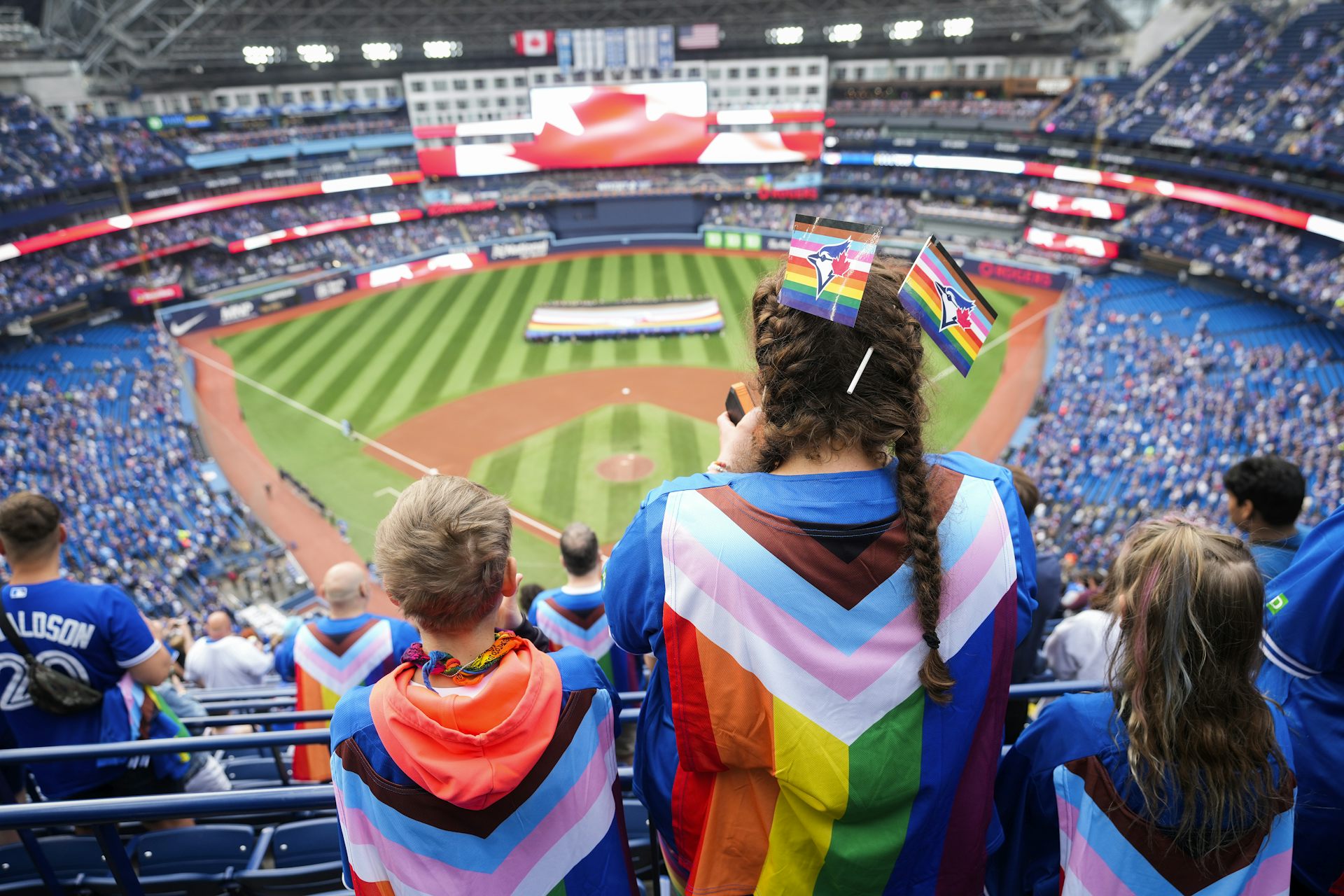 Los fanáticos con banderas del orgullo colgadas en la espalda se paran frente al campo, donde los voluntarios sostienen una gran bandera del orgullo.