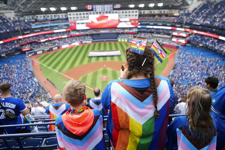 Los fanáticos con banderas del orgullo colgadas en la espalda se paran frente al campo, donde los voluntarios sostienen una gran bandera del orgullo.