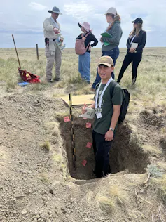 A man smiles, standing knee deep in a rectangular dug-out hole in a field with four other people standing behind.
